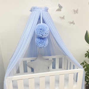 Peut inclure: Une moustiquaire de berceau bébé bleu clair avec deux gros pompons qui pendent. La moustiquaire est drapée sur un berceau blanc avec un matelas blanc et un oreiller en forme d'étoile gris. Le berceau est sur un tapis blanc.