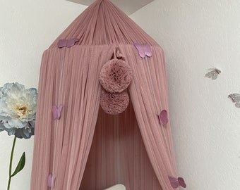 May include: A pink tulle canopy with butterfly decorations hangs over a white crib with a pink braided bumper. The canopy is decorated with two large pink pom-poms.