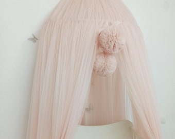 May include: A powder pink canopy with a tulle design, adorned with two pom-poms. The canopy is draped over a white crib, complemented by a braided bumper in shades of pink and white. The text "Powder Pink" is visible.