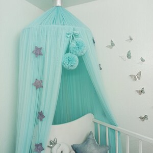 May include: A light blue canopy with silver stars and pom-poms hangs over a white crib with a white swan pillow and a silver star pillow. The wall is painted a light blue and has silver butterfly decals.