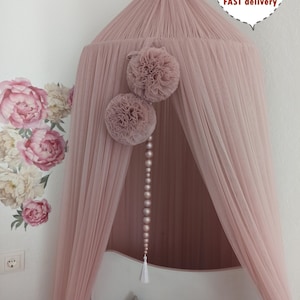 May include: A dusty pink canopy with a tulle design, adorned with two decorative pom-poms and a string of beads. The canopy is suspended from the ceiling and has a white tassel. The words "Dusty Pink" are written on the bottom right.