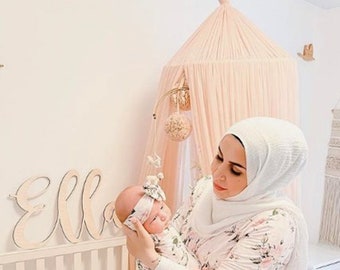 May include: A woman wearing a white floral robe and a white headscarf holds a baby wrapped in a white swaddle. The baby is wearing a headband with a bow. The woman is standing in a nursery with a white crib and a pink canopy.