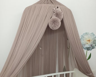 May include: A beige canopy with pom poms hangs over a white crib with a white, beige, and brown braided bumper. The crib is in a room with white walls.