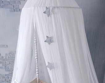 May include: White sheer canopy with silver star accents hanging from the top. The canopy is draped over a wooden crib with a white stuffed animal inside. The text "Color - Ivory" is visible in the bottom left corner of the image.