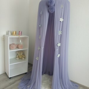 May include: A lavender tulle canopy with silver glitter stars hanging from the top. The canopy is set up in a room with a white shelf and a fluffy white rug.