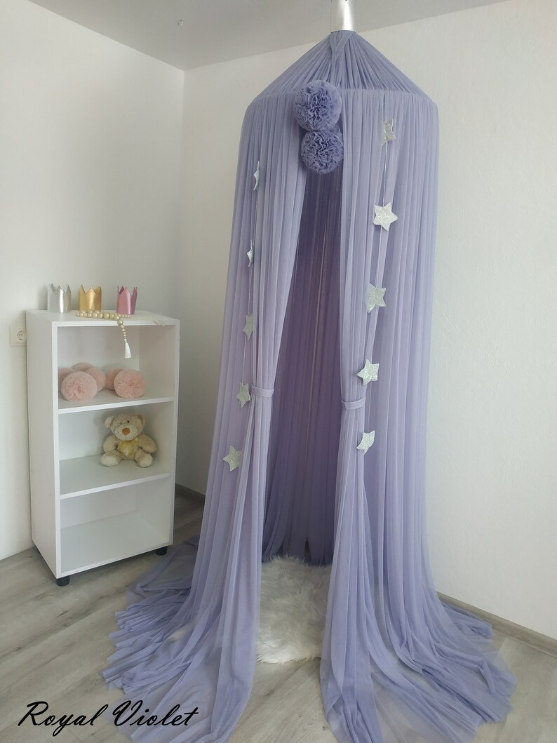 May include: A lavender tulle canopy with silver glitter stars hanging from the top. The canopy is set up in a room with a white shelf and a fluffy white rug.