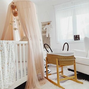 May include: A white crib with a pink canopy and a white and gray patterned blanket. A wicker bassinet on a wooden stand is in the background. The bassinet has a white liner and a white rug is on the floor. The text "26 weeks" is visible on a wooden sign.