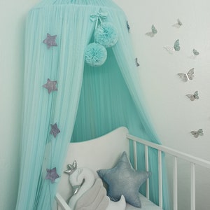 May include: A light blue canopy with silver stars and pom-poms hangs over a white crib with a silver star pillow and a white swan pillow. The wall behind the crib is light blue and has silver butterflies on it. The word "Tiffany" is written on the crib sheet.