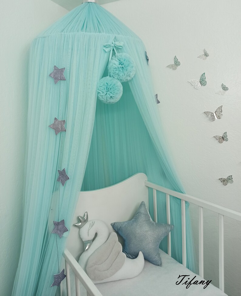 May include: A light blue canopy with silver stars and pom-poms hangs over a white crib with a silver star pillow and a white swan pillow. The wall behind the crib is light blue and has silver butterflies on it. The word "Tiffany" is written on the crib sheet.