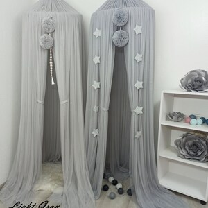 May include: Two gray tulle canopy beds with pom poms and star decorations. The bed on the left is light gray and the bed on the right is a darker gray.