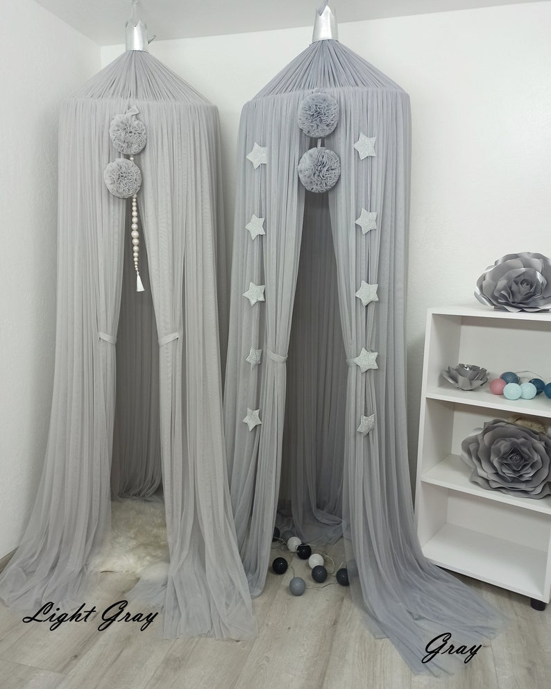 May include: Two gray tulle canopy beds with pom poms and star decorations. The bed on the left is light gray and the bed on the right is a darker gray.