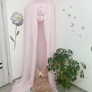 May include: A pink tulle canopy with pom poms hanging from the top. The canopy is set up in a corner of a room with a faux fur rug and a pink pillow inside.