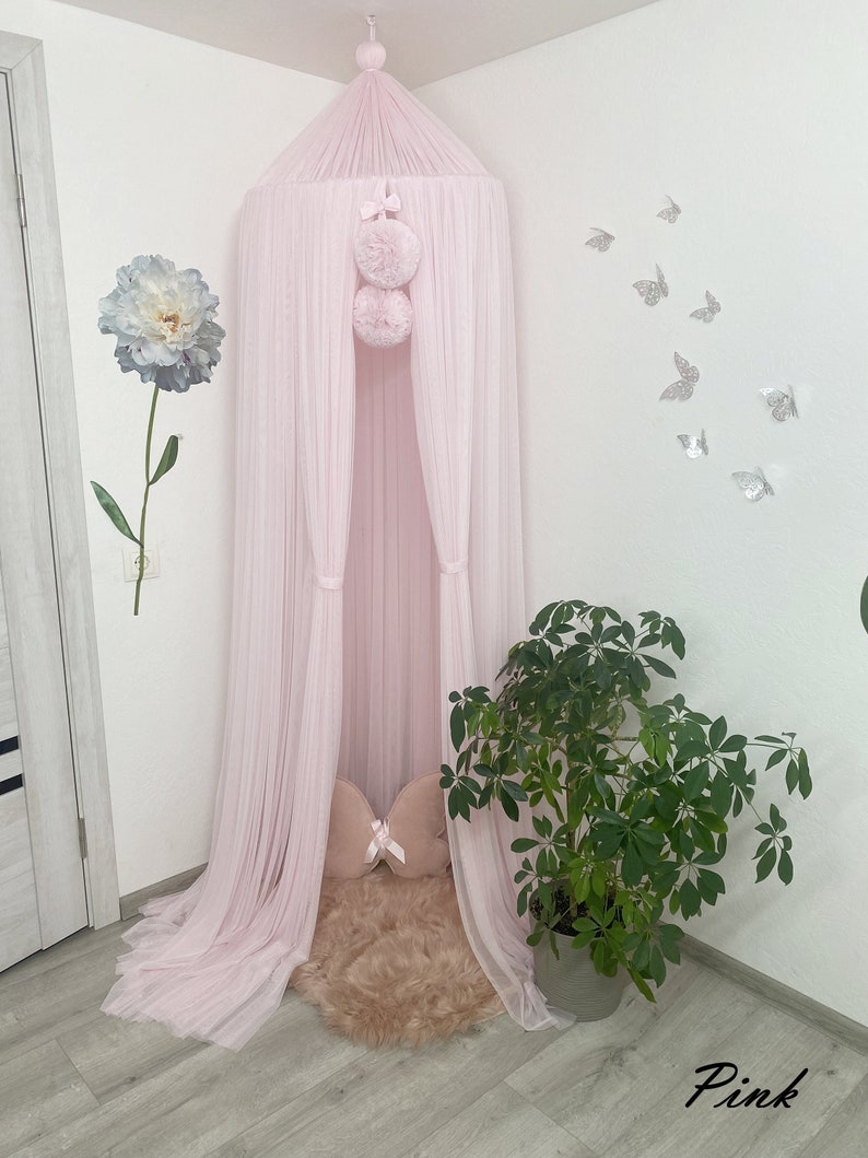 May include: A pink tulle canopy with pom poms hanging from the top. The canopy is set up in a corner of a room with a faux fur rug and a pink pillow inside.