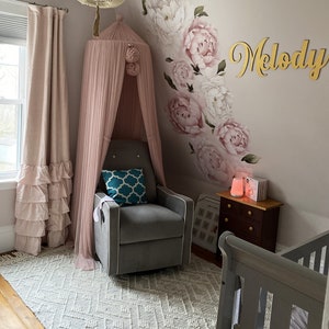 May include: A nursery room with a gray rocking chair, a pink canopy, a white and gray rug, a crib, and a wall decorated with a floral design and the name "Melody".