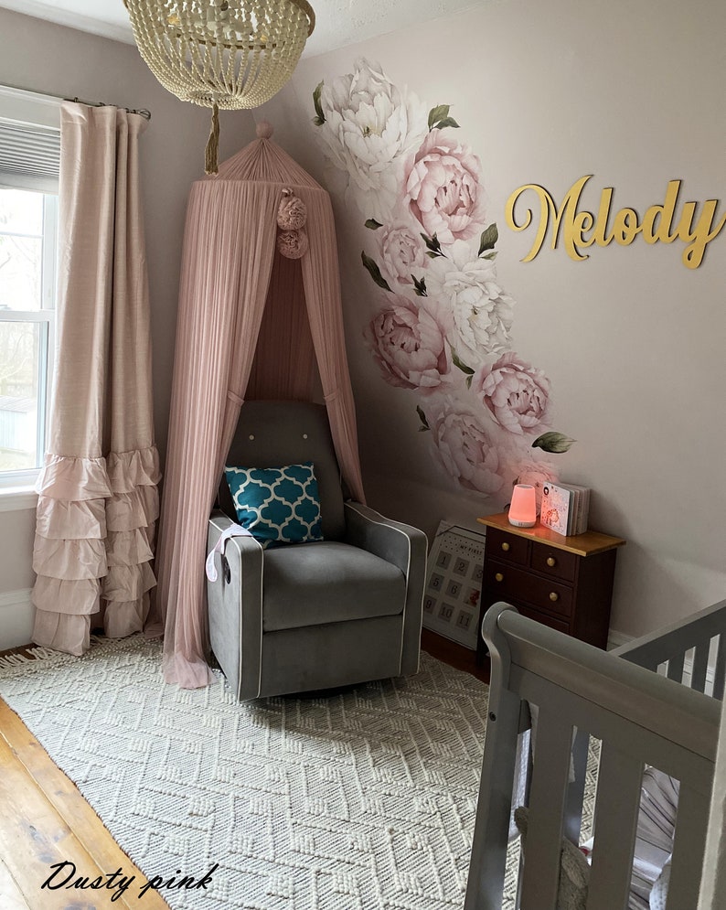 May include: A nursery room with a gray rocking chair, a pink canopy, a white and gray rug, a crib, and a wall decorated with a floral design and the name "Melody".