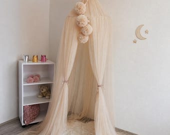 May include: A champagne-coloured tulle canopy featuring a gold crown topper and a vertical string of four matching pom-poms. The sheer fabric drapes from the ceiling to the floor, creating a soft, flowing enclosure with gathered sides.