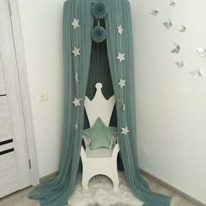 May include: A teal green canopy with white stars and pom poms hangs over a white wooden chair with a teal green star-shaped pillow. The canopy is in a corner of a room with white walls and light wood floors.
