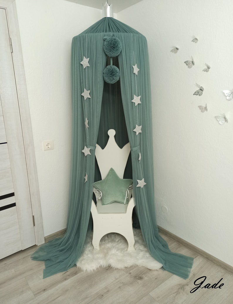 May include: A teal green canopy with white stars and pom poms hangs over a white wooden chair with a teal green star-shaped pillow. The canopy is in a corner of a room with white walls and light wood floors.