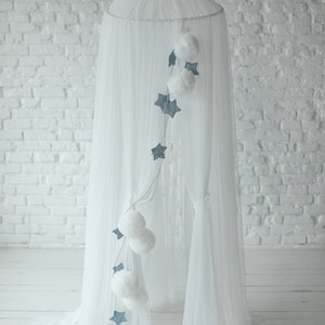 May include: White tulle canopy with a string of blue stars and white pom-poms hanging from it. The canopy is set up in a room with white brick walls.