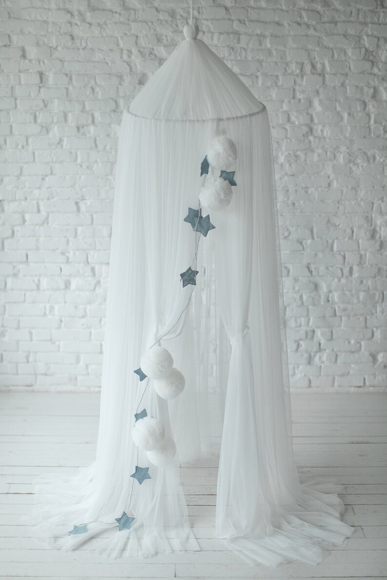 May include: White tulle canopy with a string of blue stars and white pom-poms hanging from it. The canopy is set up in a room with white brick walls.