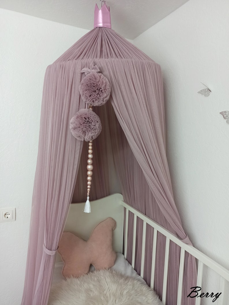 May include: A dusty rose tulle canopy with a pink crown on top. The canopy is draped over a white crib with a pink butterfly pillow. There are two pink pom poms hanging from the canopy with a beaded strand.