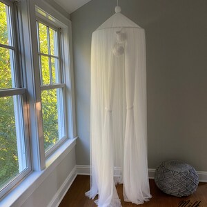 May include: White sheer canopy with white pom poms hanging from the top. The canopy is set up in a room with a gray woven footstool.