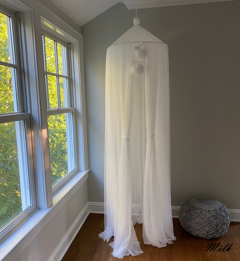 May include: White sheer canopy with white pom poms hanging from the top. The canopy is set up in a room with a gray woven footstool.