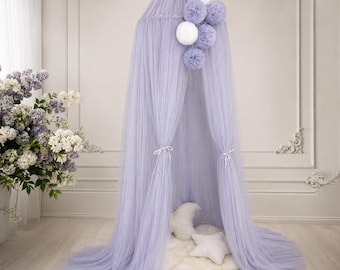 May include: A light purple tulle canopy featuring a gathered top with a cluster of white and light purple pom-poms. The sheer fabric drapes downward to the floor, with two side panels tied back. A white crescent moon and star pillow sit below.