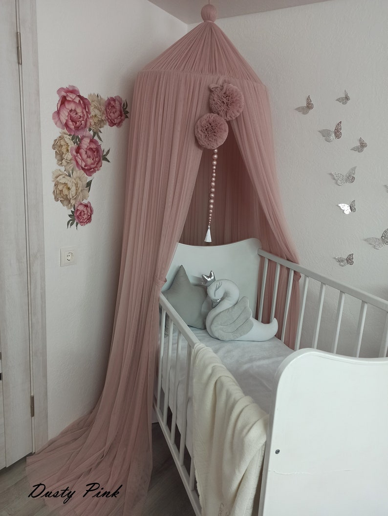 May include: A dusty pink tulle canopy with pom poms and a beaded chain hanging from the top, over a white crib with a gray swan pillow and a white blanket. The wall behind the crib has silver butterfly stickers.