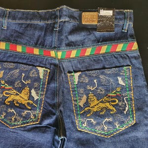 Può includere: Jeans in denim blu scuro con ricami di leoni e bandiere sulle tasche posteriori. La cintura presenta un motivo a righe rosse, verdi e oro. È visibile un'etichetta dorata.