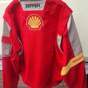 May include: A red racing jacket with tan and yellow accents. The back features the Ferrari and Shell logos. The jacket has the words "Servizio A" printed on a yellow stripe on the sleeve.