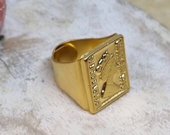 May include: A gold-colored ring with a square-shaped design featuring a profile of a woman wearing a crown. The ring is set against a light wood background.