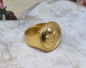 May include: A gold-toned ring with a round, raised design featuring a fleur-de-lis. The ring is on a white wooden surface.