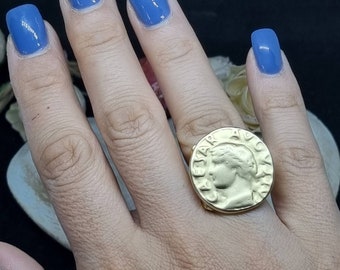 May include: A gold-toned ring featuring a large, round coin design with a profile of a person's face and text. The ring is worn on a person's finger.
