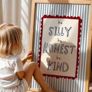May include: A rectangular wooden framed wall art piece featuring a light blue and white vertical striped background. A central cream-colored rectangle with a scalloped red border contains the dark blue text: be SILLY be HONEST be KIND.