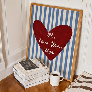 May include: Framed artwork featuring a red heart with the text "Ok, love You, Bye" on a blue and white striped background. A stack of books and a white mug with coffee are in front of the artwork.