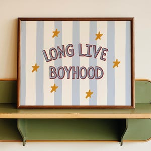 Może przedstawiać: Oprawiona grafika z napisem "LONG LIVE BOYHOOD" w czerwonych i fioletowych literach, na tle niebiesko-białych pionowych pasków i złotych gwiazd. Grafika jest eksponowana na zielono-beżowej półce.