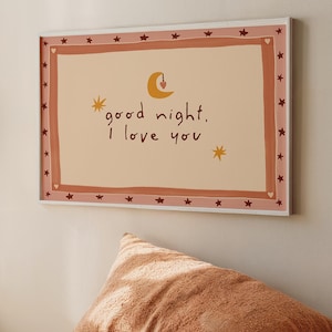 May include: A framed art print with the text "good night, I love you" in a whimsical font, surrounded by a border of stars and hearts. Two plush, orange-brown pillows are stacked below the artwork, creating a cozy, inviting scene.