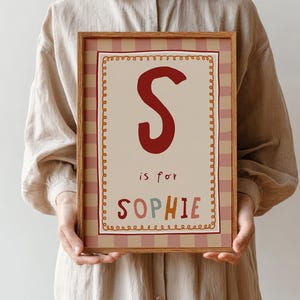 Op de afbeelding: Een rechthoekige ingelijste print met een grote donkerrode letter S, de tekst is for en de naam SOPHIE in veelkleurige letters. Het ontwerp bevat een crèmekleurig midden met een decoratieve rand en een roze en crèmekleurige gestreepte achtergrond.