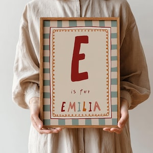 May include: A framed print featuring the letter "E" in a bold, burgundy color. The print includes the text "is for EMILIA" in a playful font. The artwork is set against a cream background with a decorative border and a light brown wooden frame.