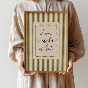 Op de afbeelding: Een rechthoekige houten lijst met een print met een achtergrond van verticale olijfgroene en crèmekleurige strepen. In het midden staat een crèmekleurige rechthoek met een geschulpte bruine rand en de zwarte tekst I am a child of God.