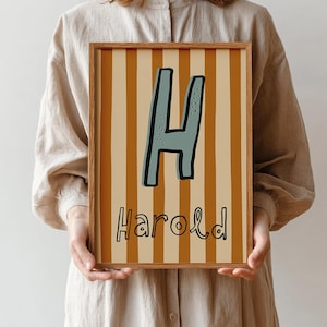 May include: A framed print with the name "Harold" in a playful font. The letter "H" is teal with a black outline, set against vertical tan and cream stripes. The frame is light brown. A decorative piece for a child's room.