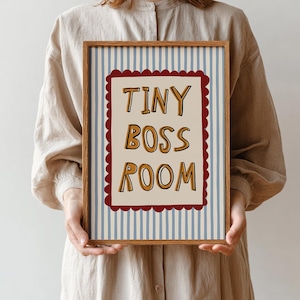 May include: A framed print with the words "TINY BOSS ROOM" in a playful font. The artwork features a cream-colored background with the text in brown and yellow, framed by a red scalloped border. The background has blue and white vertical stripes.