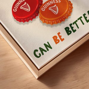 May include: A framed canvas print featuring two Guinness bottle caps, one red and one orange, with the Guinness logo. The phrase "CAN BE BETTER" is printed in green and orange. The frame is made of light-colored wood.