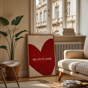 May include: A framed art print featuring a large red heart with the words "THE ART OF LOVING" in white. The artwork is displayed in a living room setting with a beige sofa, wooden chair, and a potted plant. The room has a window with a view of a building.