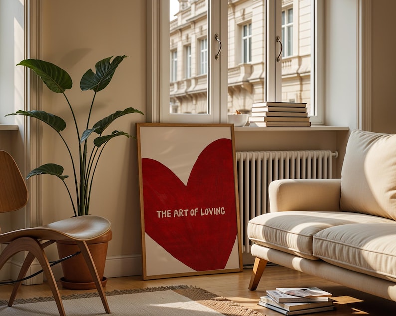 May include: A framed art print featuring a large red heart with the words "THE ART OF LOVING" in white. The artwork is displayed in a living room setting with a beige sofa, wooden chair, and a potted plant. The room has a window with a view of a building.
