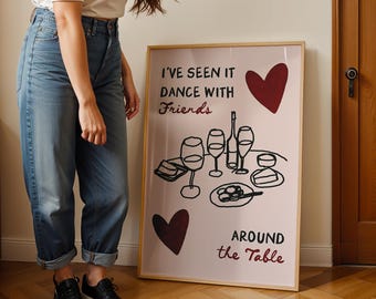 Puede incluir: Una impresión artística enmarcada con un fondo rosa claro presenta el texto "I've seen it dance with Friends around the Table". La obra de arte incluye ilustraciones de copas de vino, una botella y comida, con dos corazones burdeos.