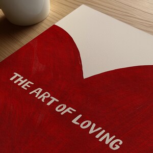 May include: A red and white art print with the text "THE ART OF LOVING" in white. The print has a heart-shaped design in red paint. A white mug is visible in the upper left corner, resting on a wooden surface.