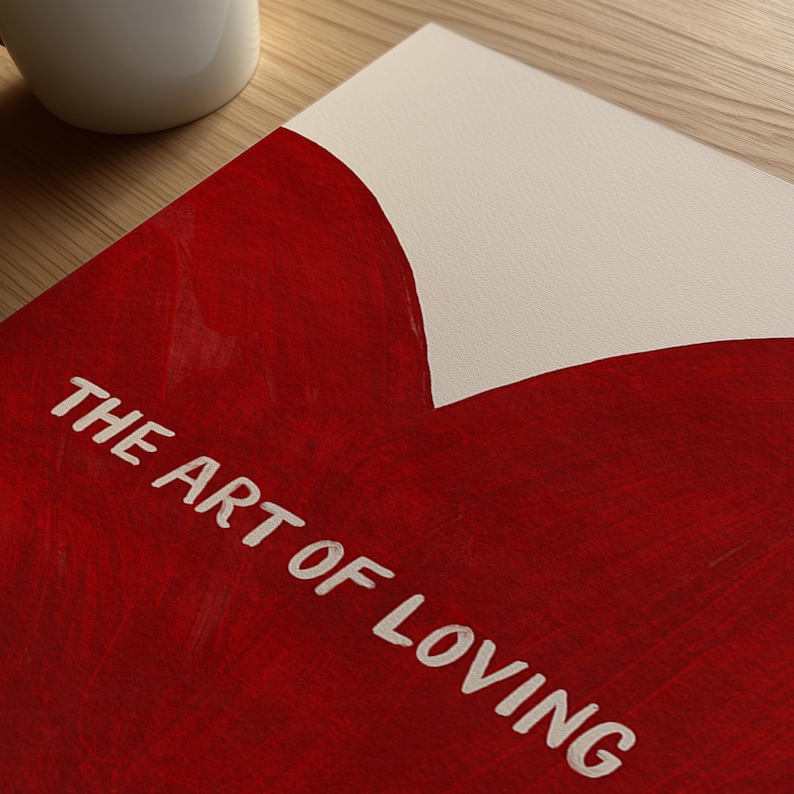 May include: A red and white art print with the text "THE ART OF LOVING" in white. The print has a heart-shaped design in red paint. A white mug is visible in the upper left corner, resting on a wooden surface.