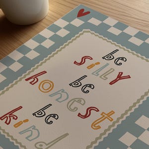 Pode incluir: Impress&atilde;o decorativa com as palavras "be silly, be honest, be kind" em letras coloridas. A impress&atilde;o tem uma borda xadrez azul claro e branco e um cora&ccedil;&atilde;o vermelho. Uma caneca branca est&aacute; no fundo.